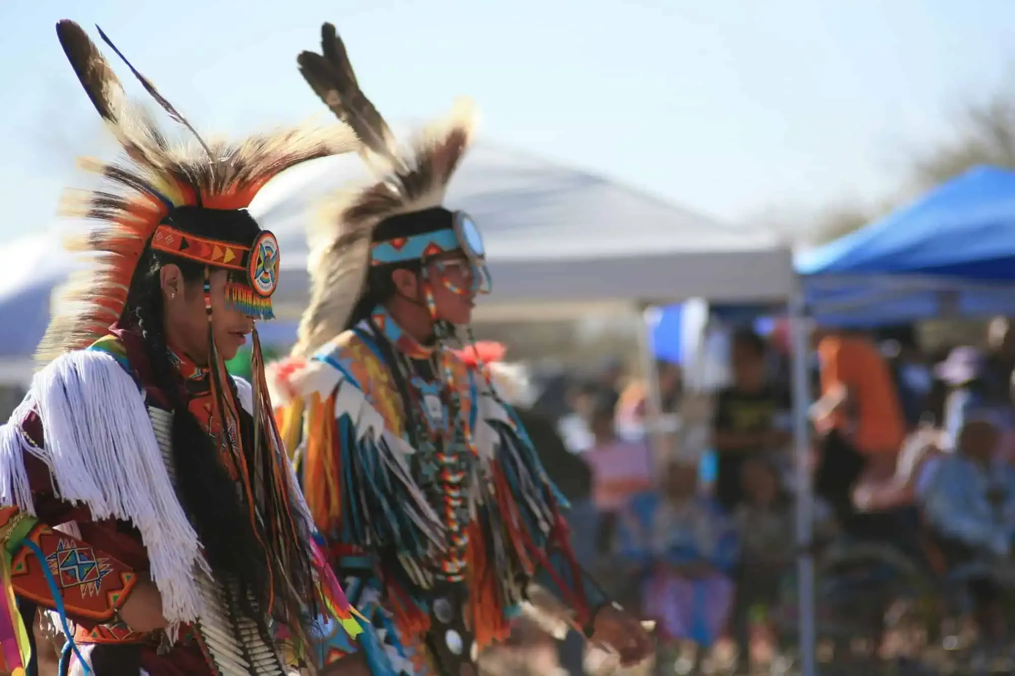 Two Native American men in colorful regalia with feathered headdresses at an outdoor gathering.