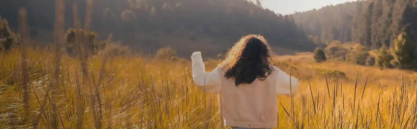 Person with curly hair walking through a sunlit field of tall grass with hills and trees in background.