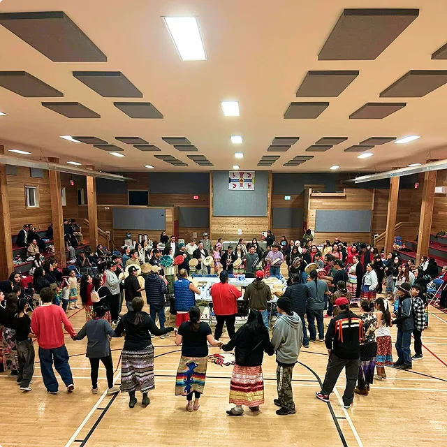 Large group holding hands in a gymnasium circle, some wearing traditional clothing and drums in the center.