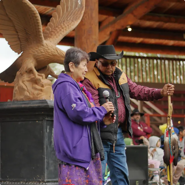 An elderly woman in purple speaks into a microphone held by a man in a black hat and vest.