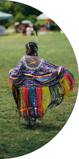 Person dancing outdoors in colorful traditional Native American regalia with long fringe and feather.
