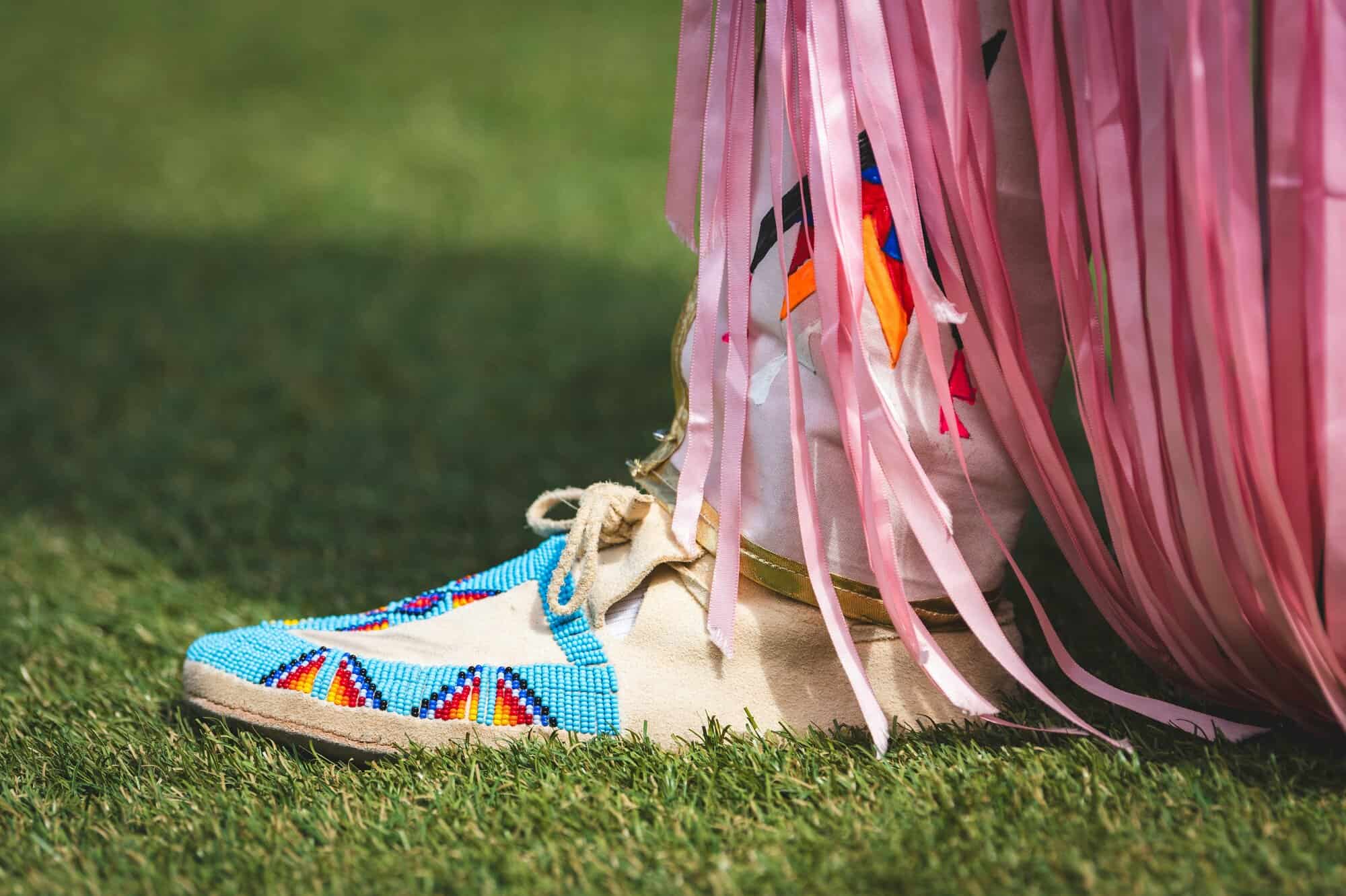 Beaded moccasin shoe with blue and colorful patterns worn with pink fringed garment on grass.