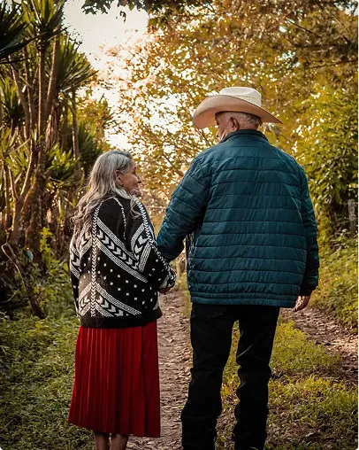 Elderly couple holding hands walking on a forest path surrounded by autumn foliage.