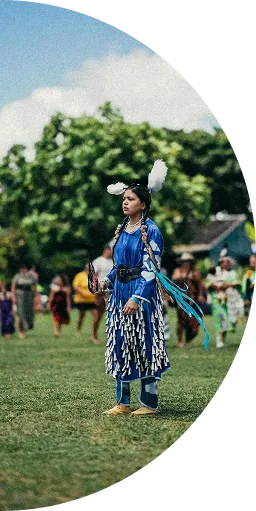 Woman in traditional blue Native American regalia standing on grass at outdoor gathering.