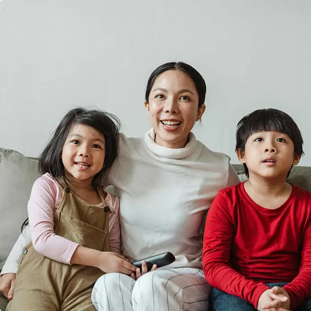 Smiling woman sitting on a couch with two children, one holding a remote control.