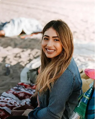 Smiling woman with long hair wearing a denim jacket sitting on a blanket at the beach.