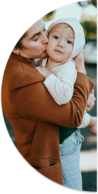 Woman in brown coat kissing a toddler wearing a white hooded outfit outdoors.