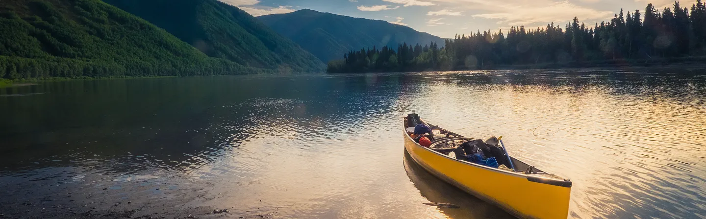 Yellow canoe floating on calm water near forested mountains at sunset.