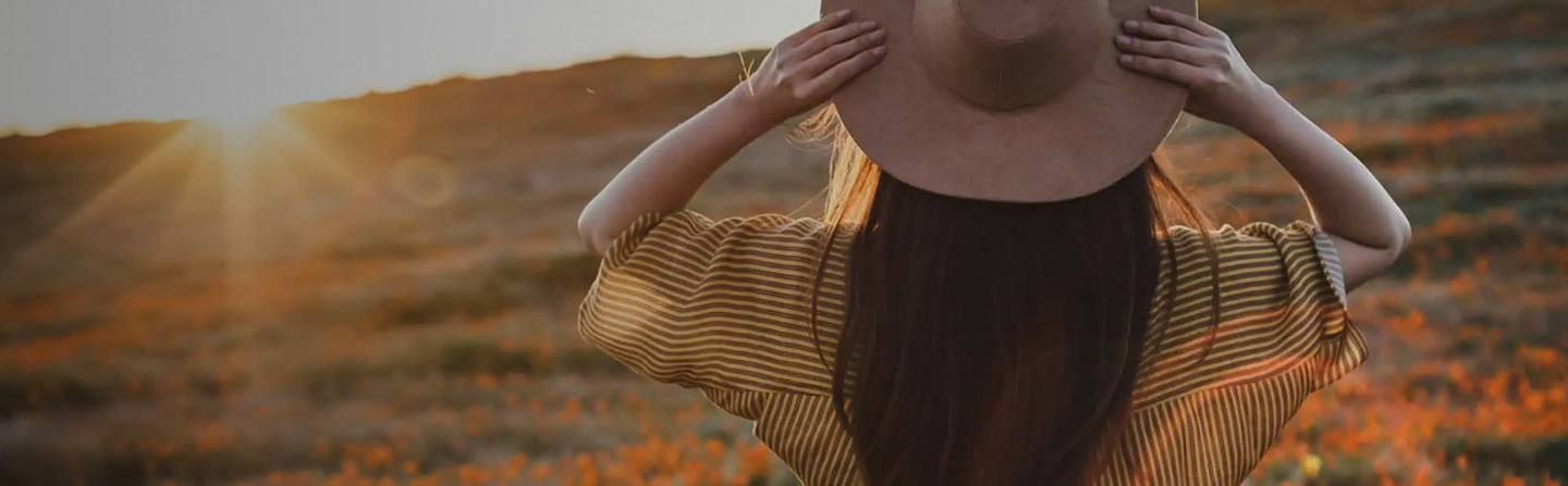 Person with long hair wearing a hat and striped shirt watches the sunset over a field with orange flowers.