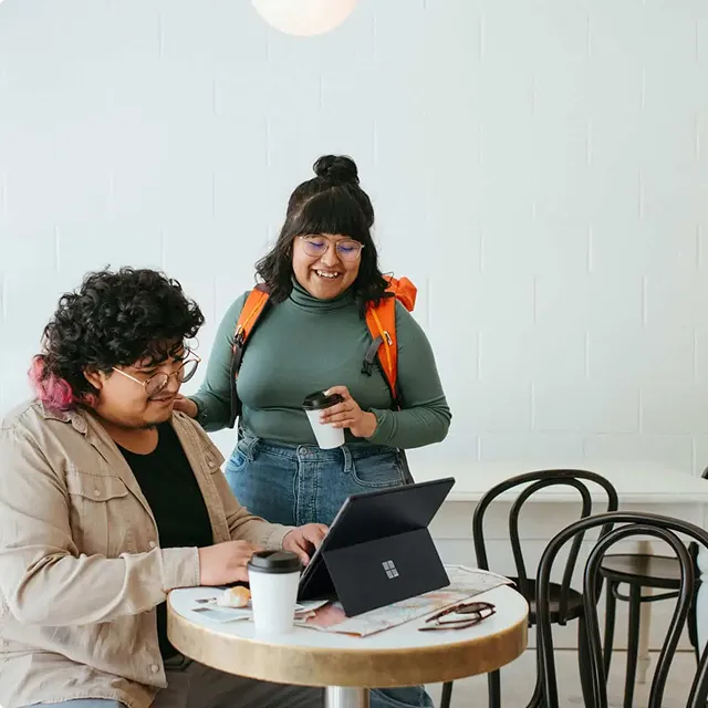 Two people share coffee while one works on a tablet at a round café table.
