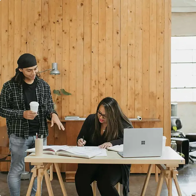 Two people working together at a desk with an open book and laptop in a wooden paneled room.