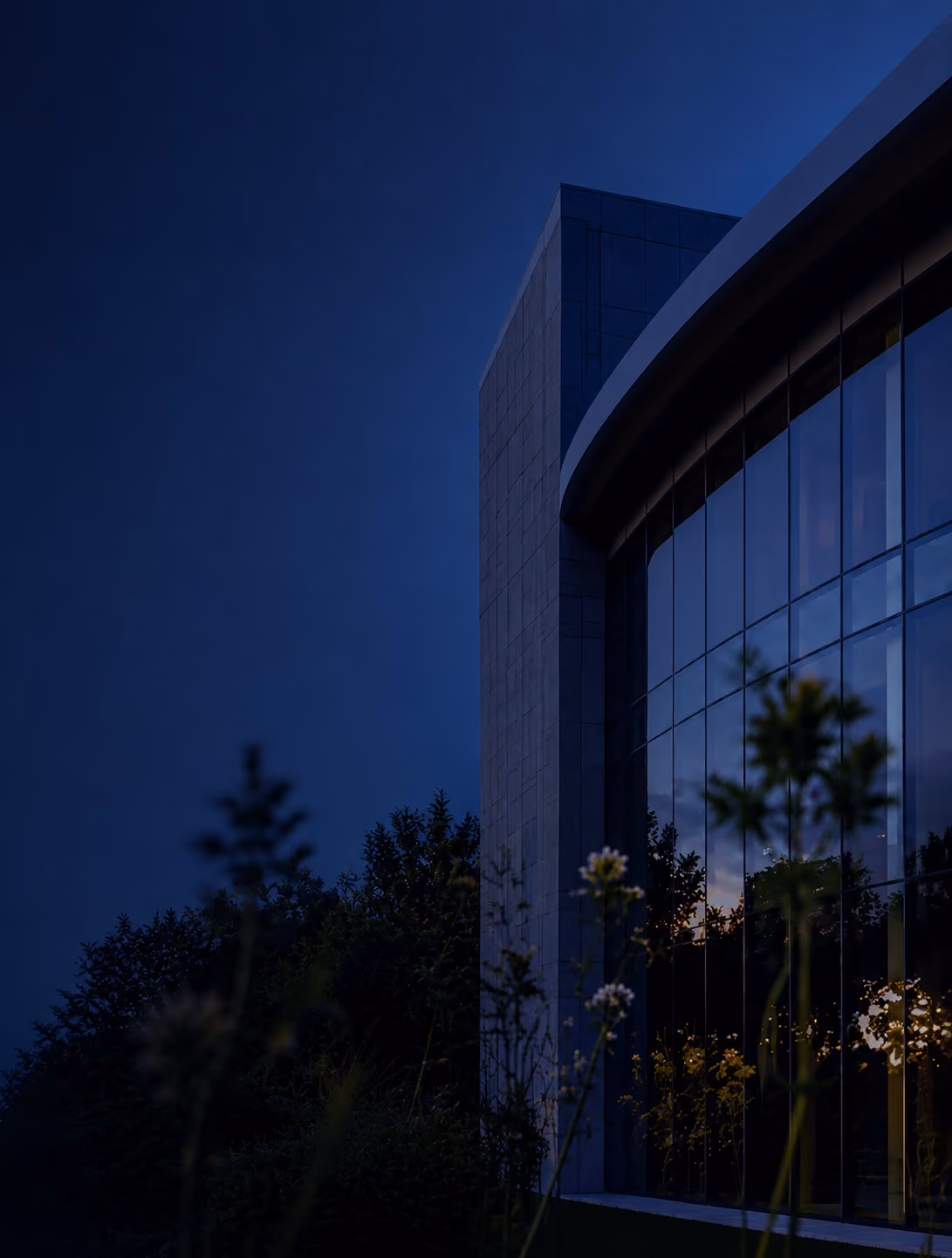 Modern building exterior with large windows and plants at dusk or dawn in natural setting outdoors.