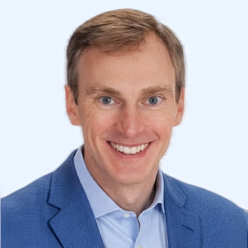 Smiling man with light brown hair wearing a blue blazer and light blue shirt on a light background.
