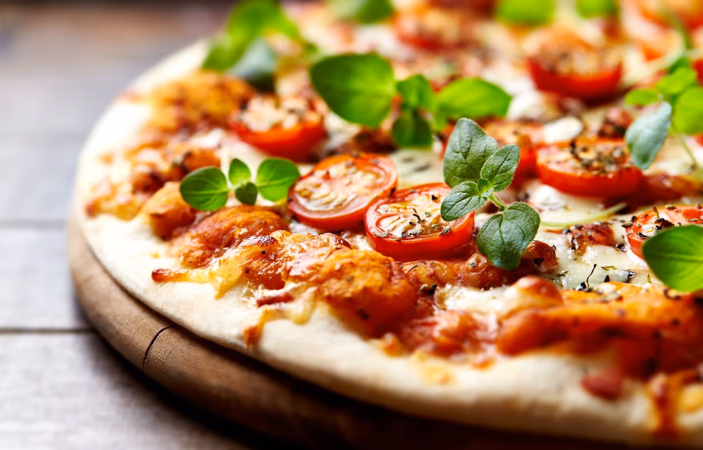 Close-up of pizza with melted cheese, tomato slices, and fresh green herbs on wooden board.