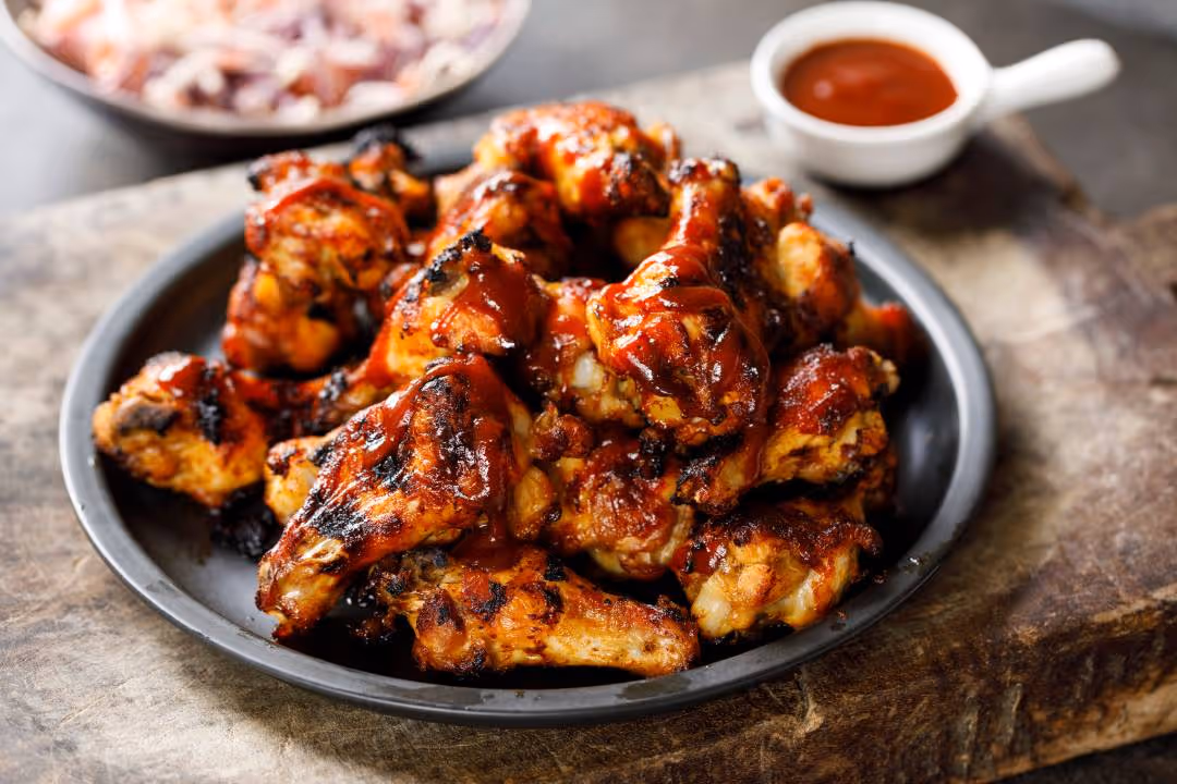 Plate of grilled chicken wings glazed with barbecue sauce on a wooden surface with sauce cup nearby.