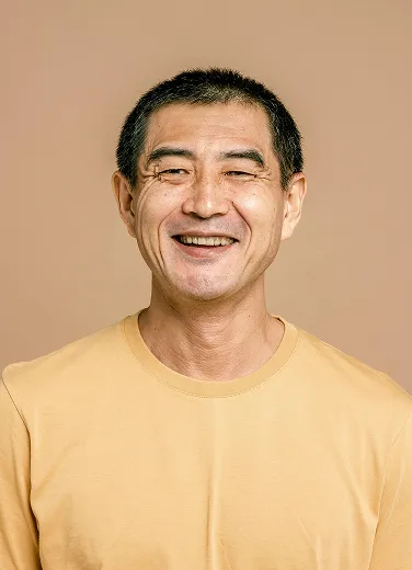 Smiling middle-aged Asian man wearing a yellow shirt against a beige background.