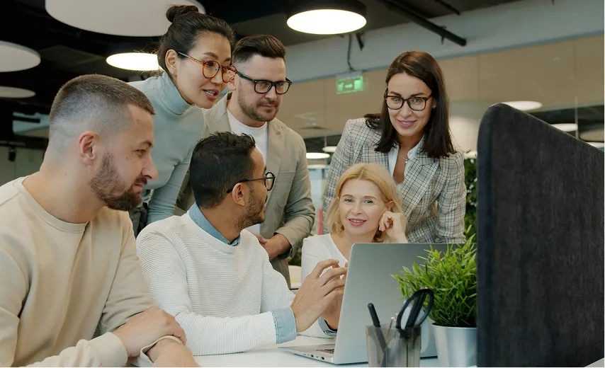 Diverse group of six people gathered around a laptop in a modern office, engaged in discussion.