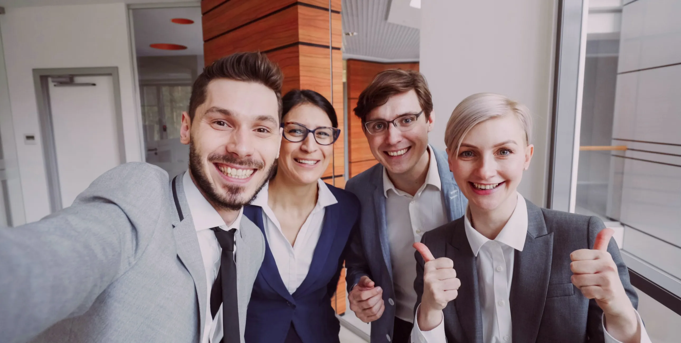 Four businesspeople in suits smiling, one giving two thumbs up in a modern office hallway.