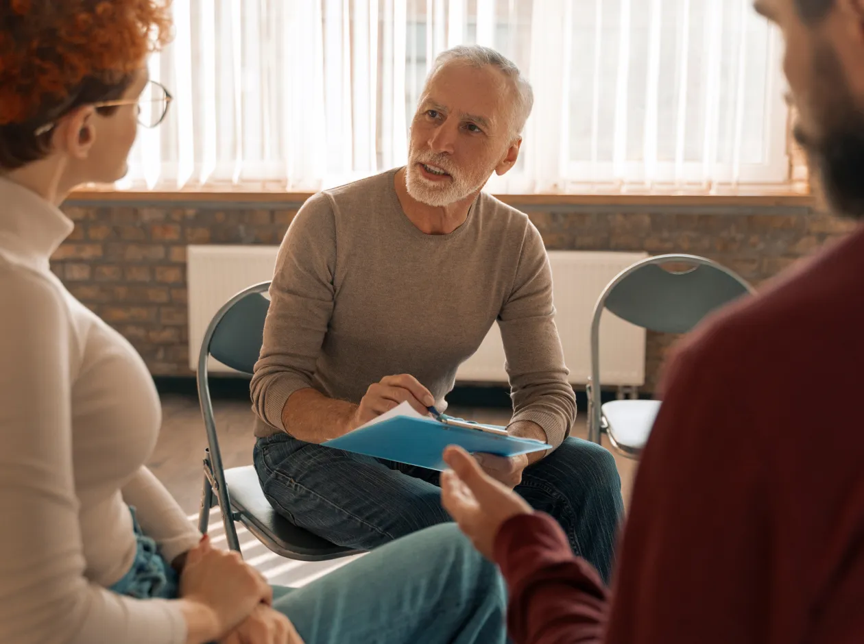 Mature man with clipboard talking to a seated man and woman in a bright room.