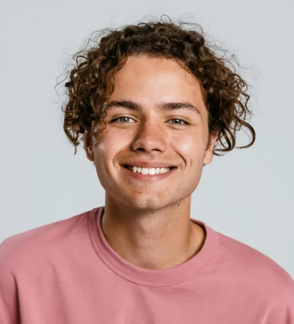 Young man with curly hair smiling and wearing a pink sweatshirt against a plain background.