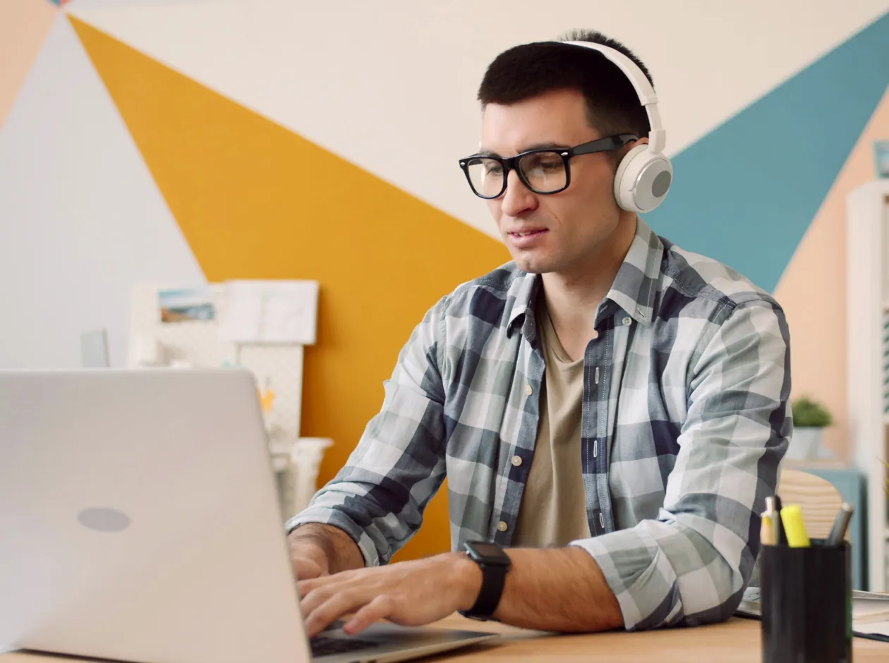 Young man wearing glasses and headphones working on a laptop at a desk indoors with colorful geometric wall art.