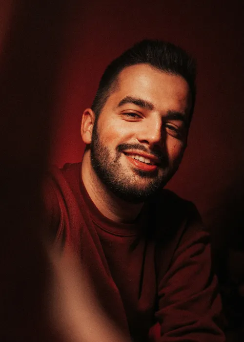 Smiling man with a beard wearing a dark red sweater against a dark red background.