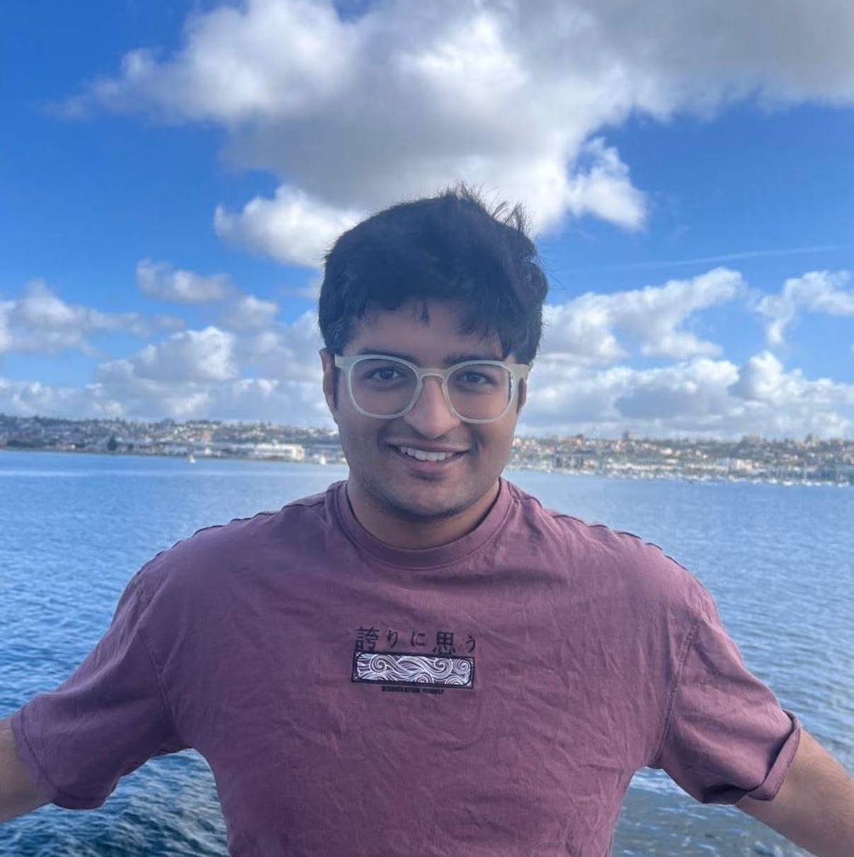 Smiling young Shivam Pujara, Founder of Luxen Talent and Madhupa standing by calm water with a cloudy blue sky.