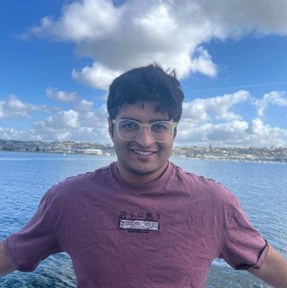 Smiling young Shivam Pujara, Founder of Luxen Talent and Madhupa standing by calm water with a cloudy blue sky.
