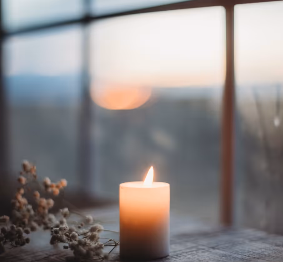 Lit candle on a table near window with blurred soft sunset and dried flowers.