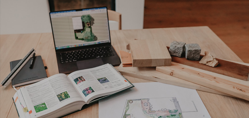 Laptop mit Gartenplan auf dem Bildschirm, auf einem Holztisch liegen ein aufgeschlagenes Buch, Gartenpläne auf Papier, Holzstücke und Steine