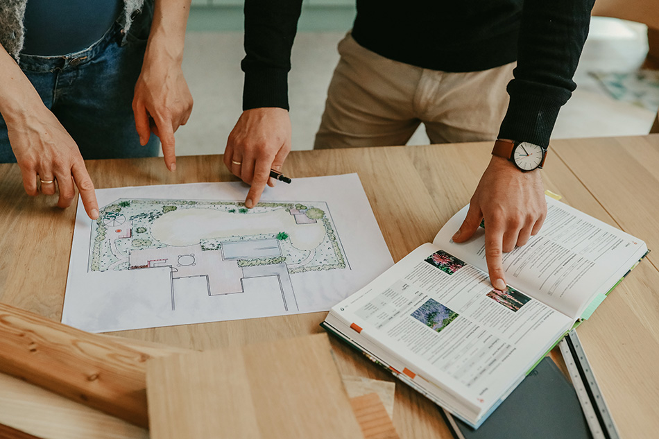 Zwei Personen zeigen mit den Fingern auf einen Gartenplan und ein aufgeschlagenes Buch auf einem Holztisch