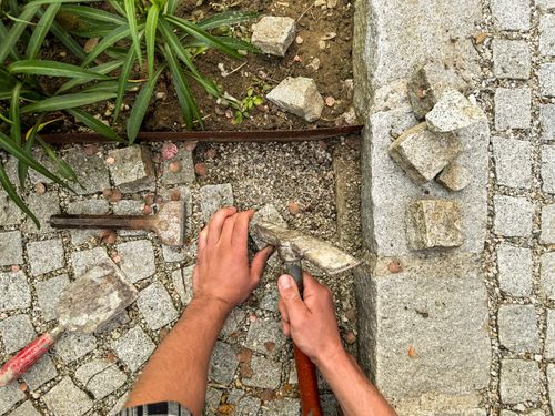 Hände, die mit Hammer und Stein Pflastersteine zwischen Rasenkanten verlegen