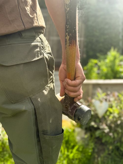 Hand hält einen Hammer mit abgenutztem Holzstiel neben einer Hose aus robustem Stoff im Freien
