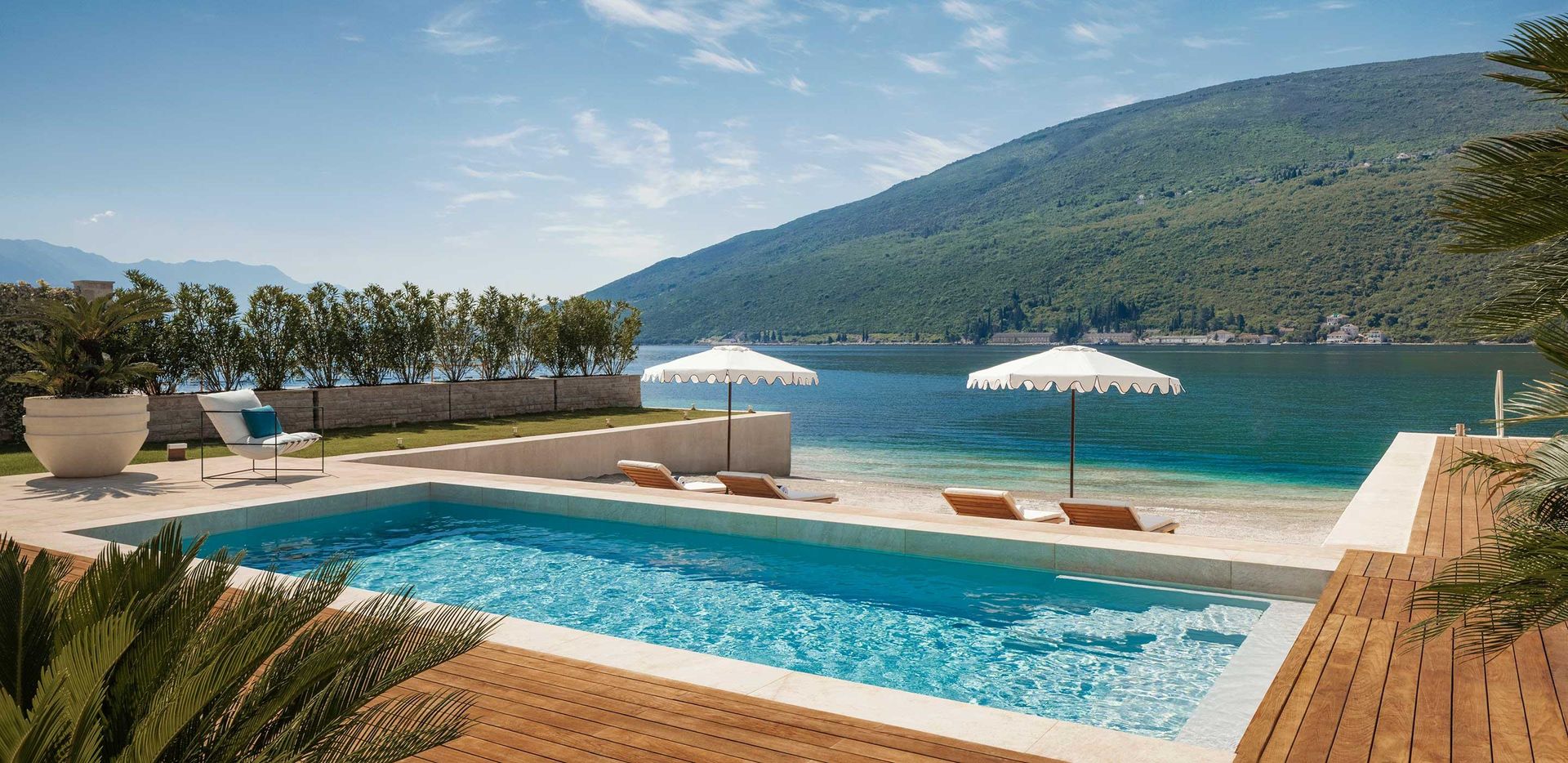 Pool on terrace with lounge chairs, umbrellas, overlooking calm blue sea and green hills under clear sky. One & Only Villa Portonovi.