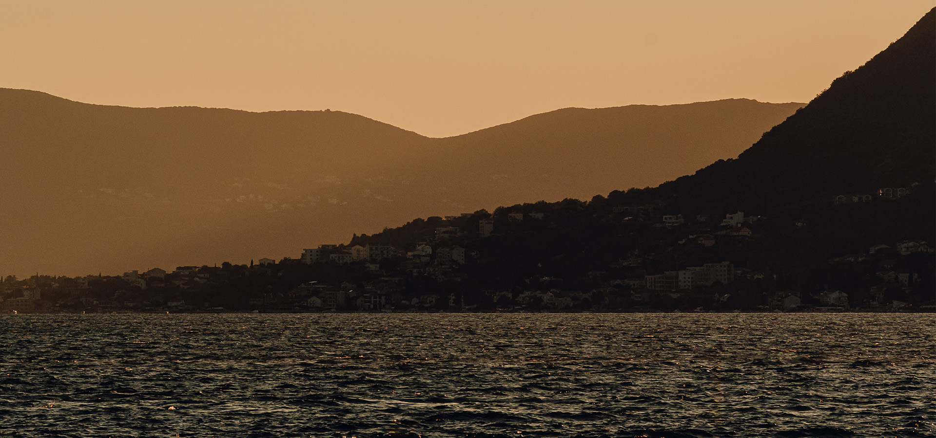 Calm sea with silhouetted coastal hillside of Boka Bay Tivat Montenegro and buildings under an orange sunset sky.