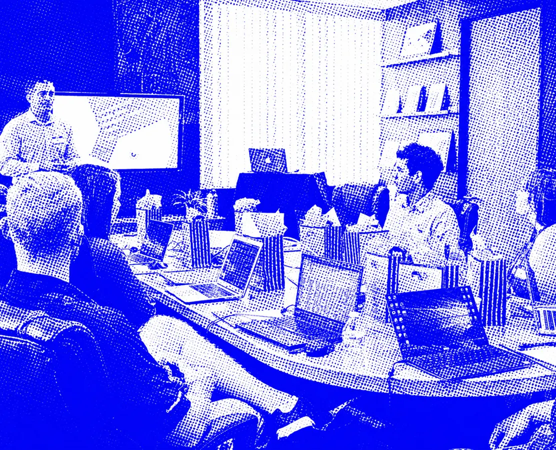 People sit around a table with laptops as one person stands and presents in a meeting room.
