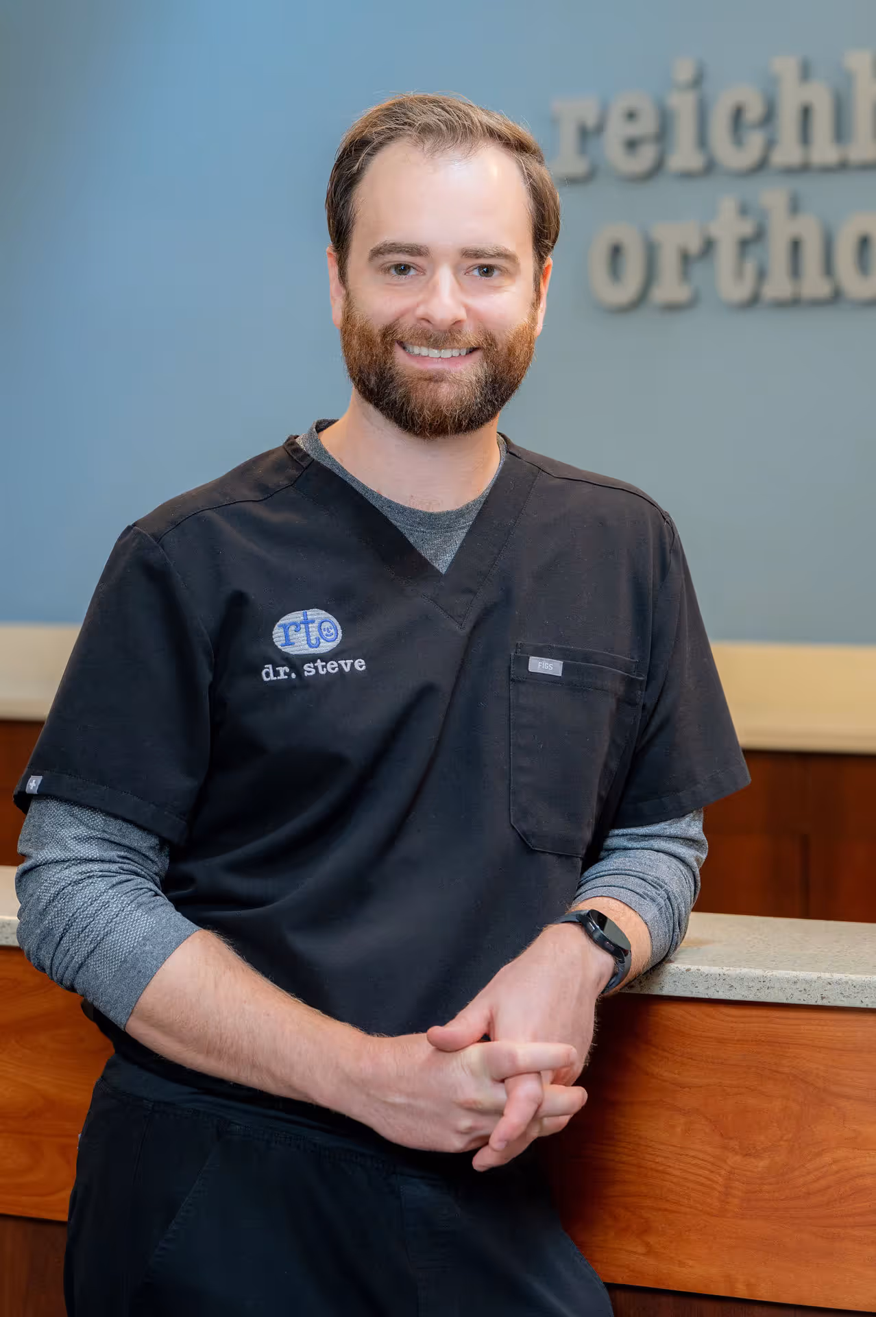Smiling male healthcare professional in black scrubs leaning on a reception desk.