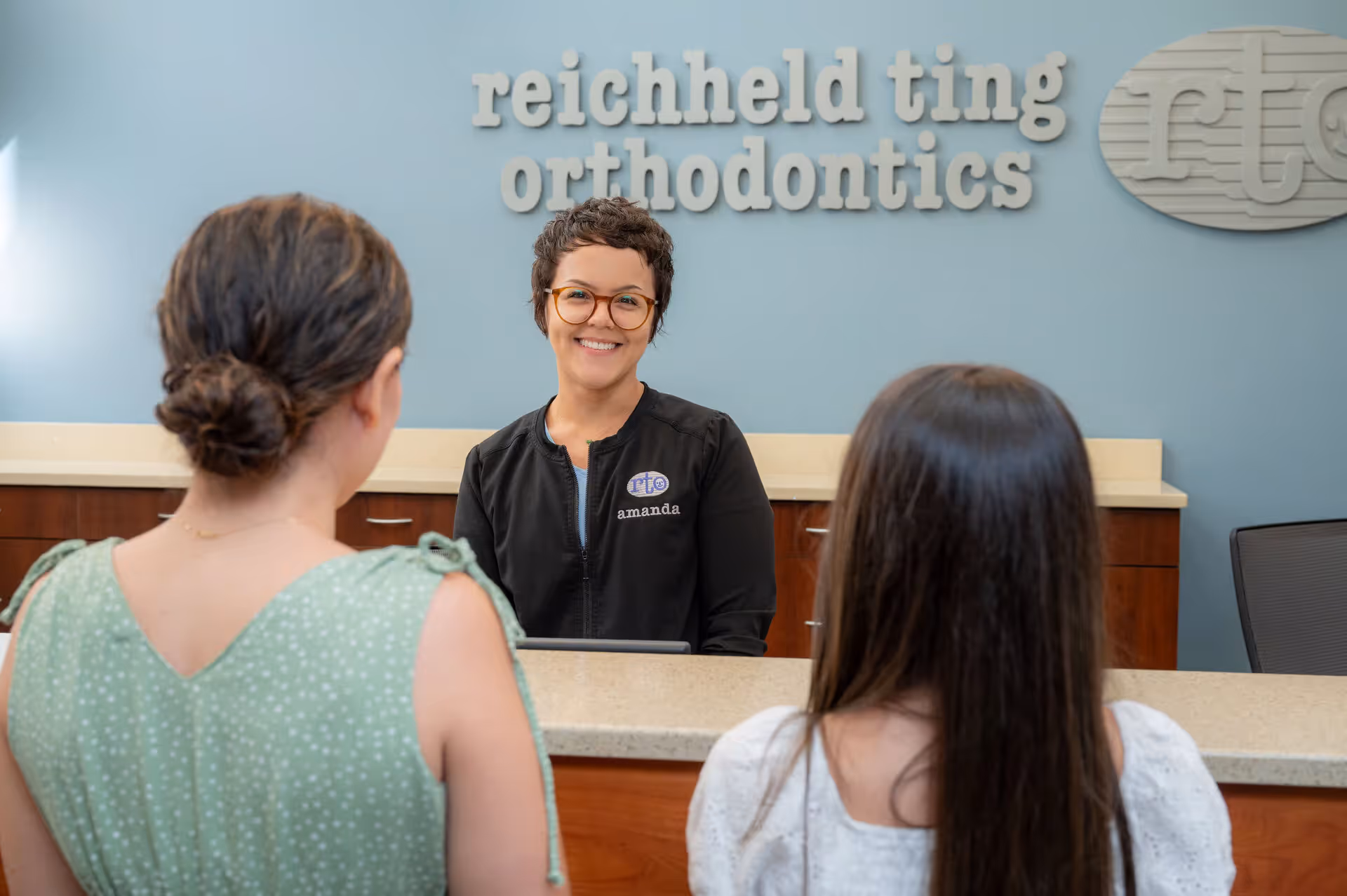 Receptionist named Amanda at orthodontics office smiles at two women standing at the counter.