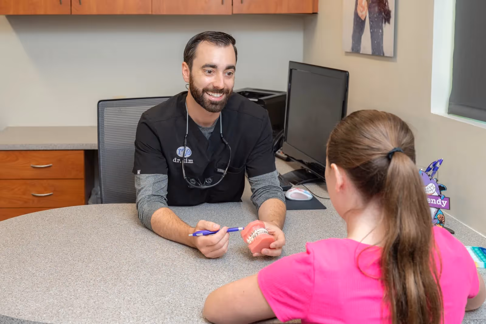 Dentist explaining braces on a dental model to a girl in a pink shirt at a clinic desk.