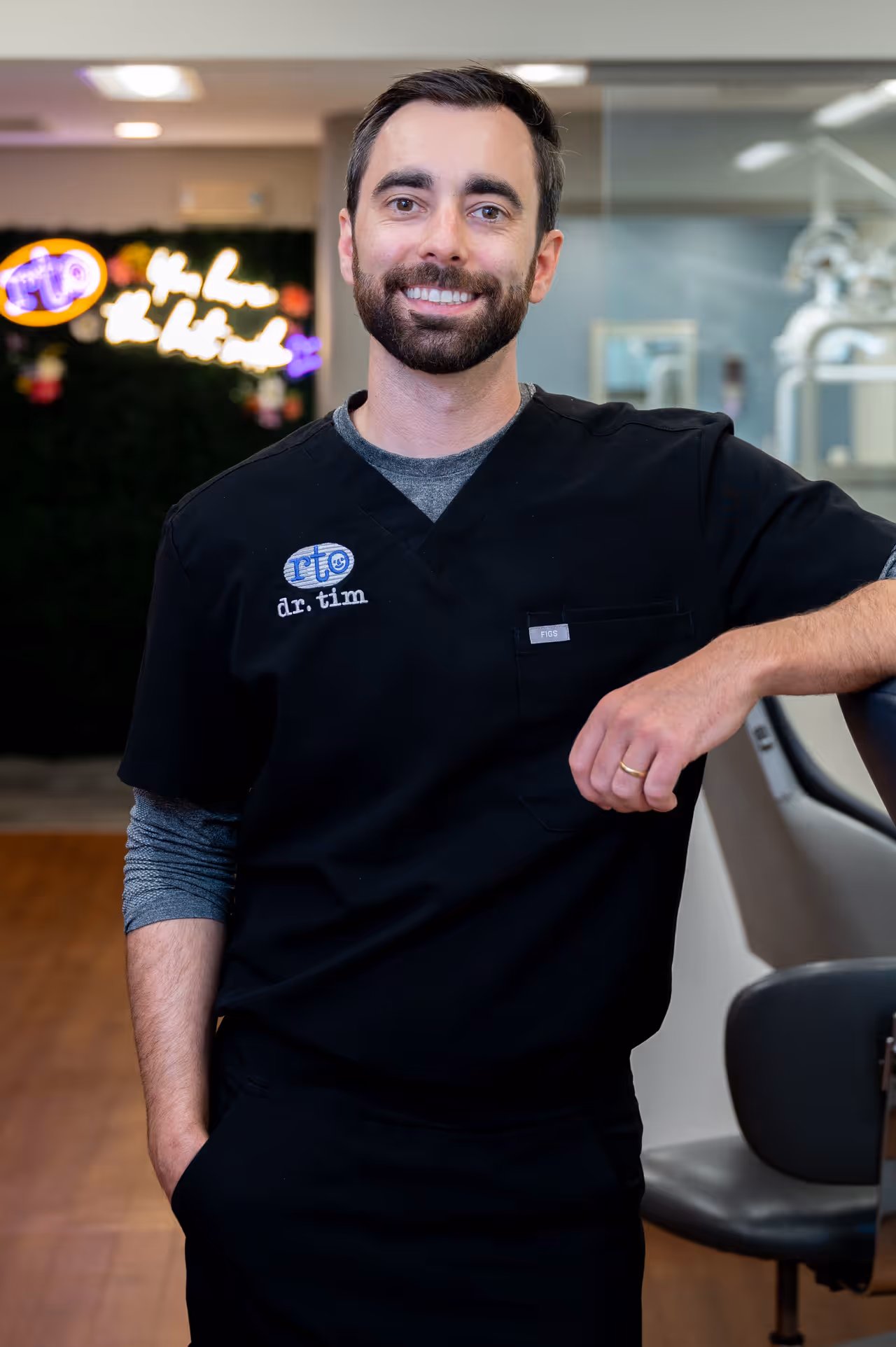 Smiling male dentist wearing black scrubs labeled 'dr. tim' standing in dental office.