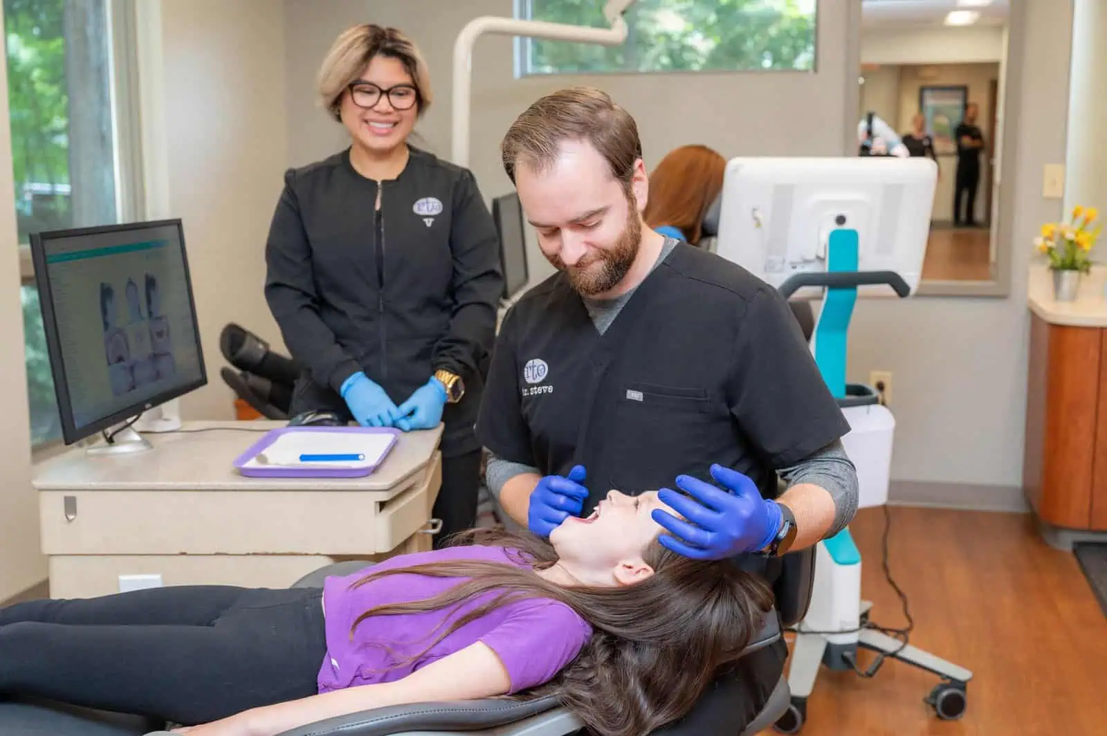 Dentist with blue gloves examining a young girl’s mouth while a dental assistant watches and smiles.