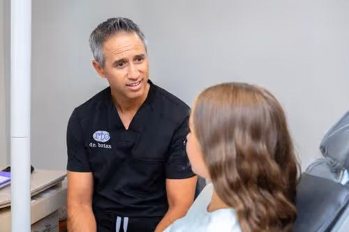 Orthodontist Dr. Brian in black scrubs talking to a seated female patient in a consultation room.