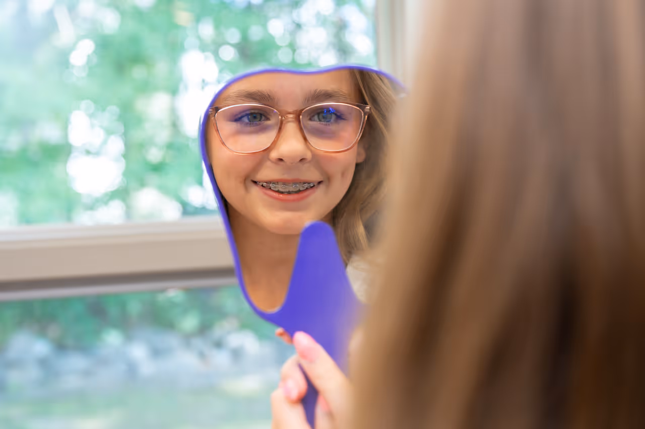 Smiling girl with glasses and braces looking at her reflection in a handheld mirror.