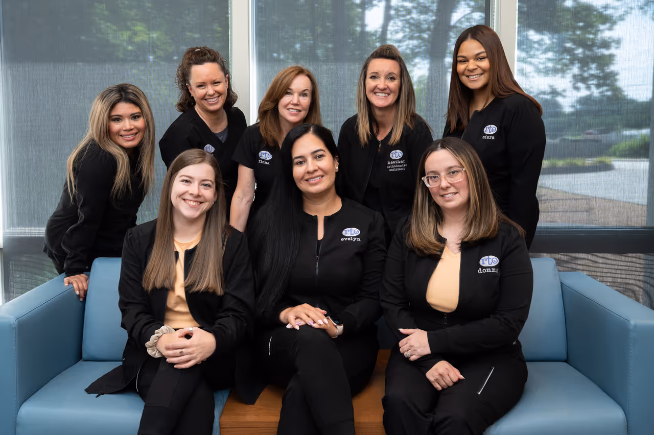 Eight smiling women in black uniforms seated and standing inside an office with large windows behind.