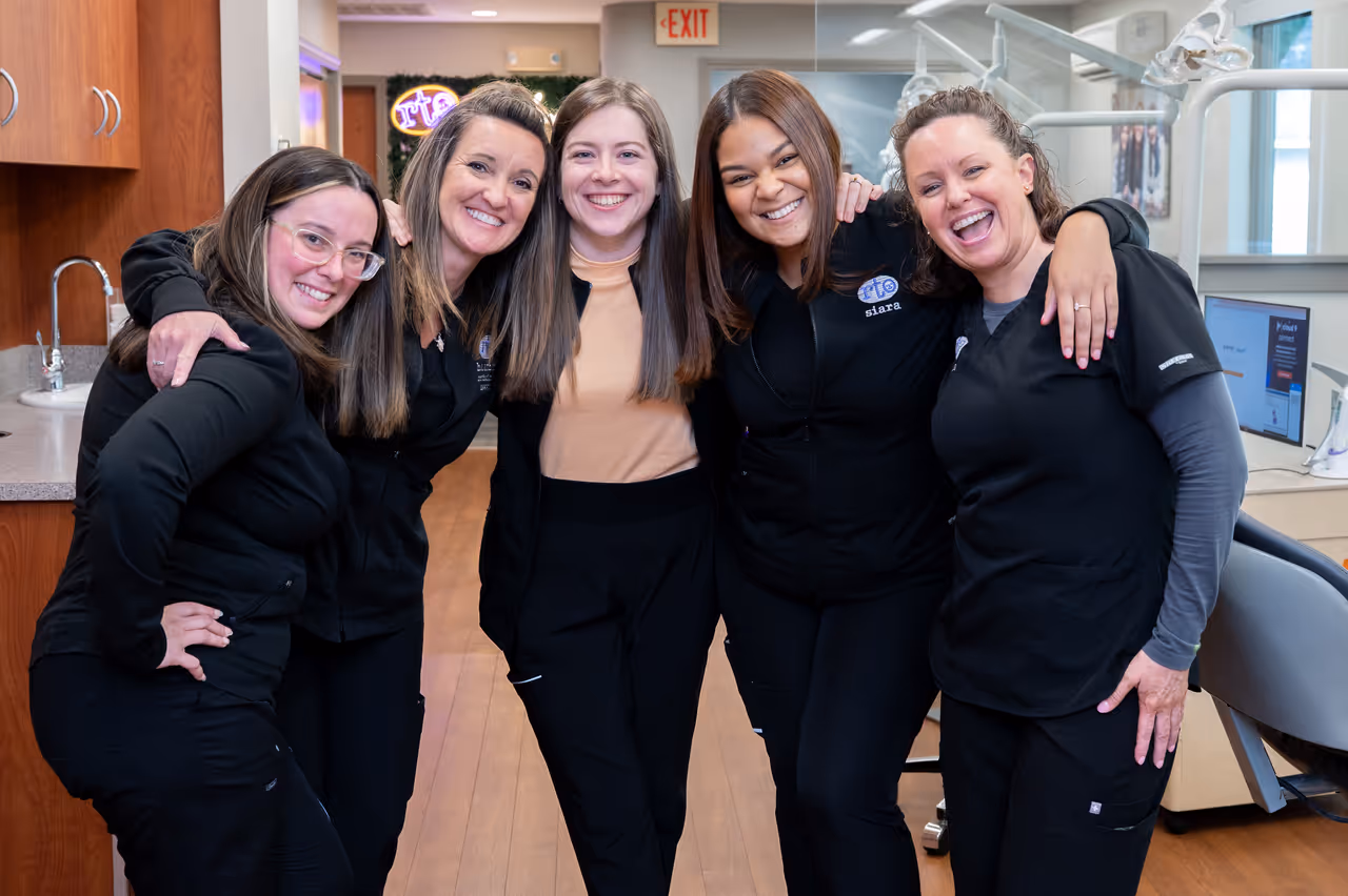Five women smiling together in a dental office wearing black scrubs and casual clothes.