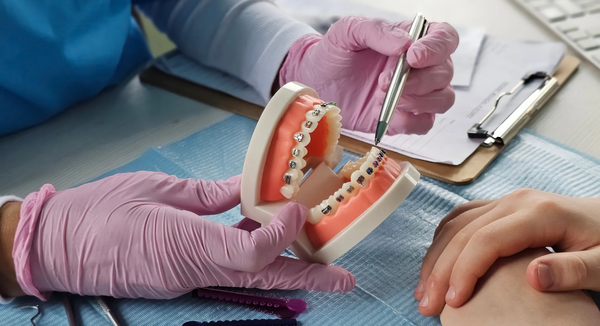 Dentist wearing pink gloves showing teeth model with braces while a patient watches.