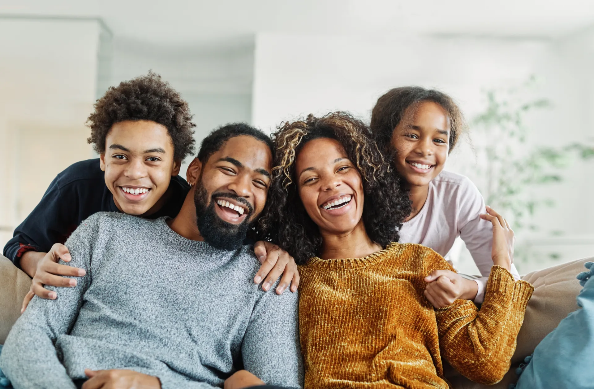 Smiling family of four sitting closely together on a couch in a bright living room.