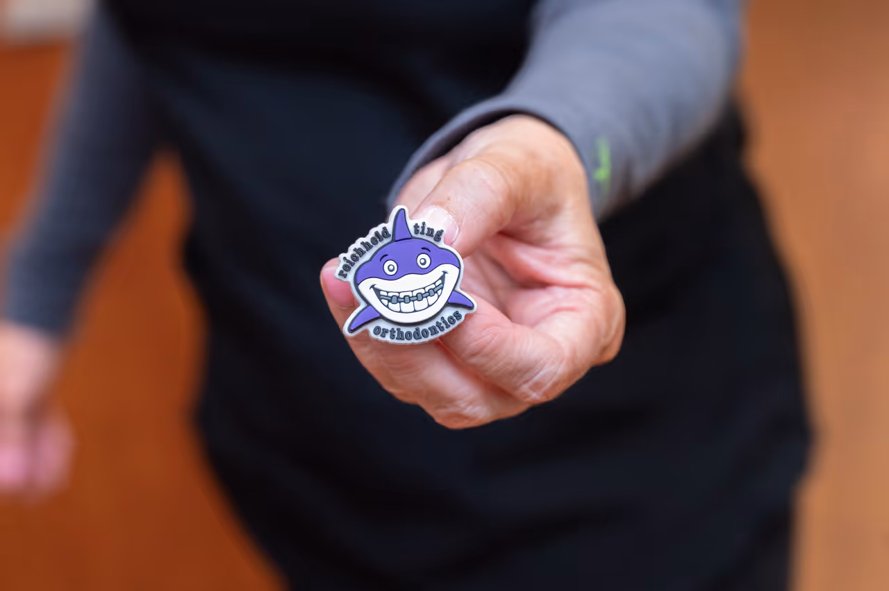 Hand holding a purple shark sticker with braces and text reading Reichheld Ting Orthodontics.