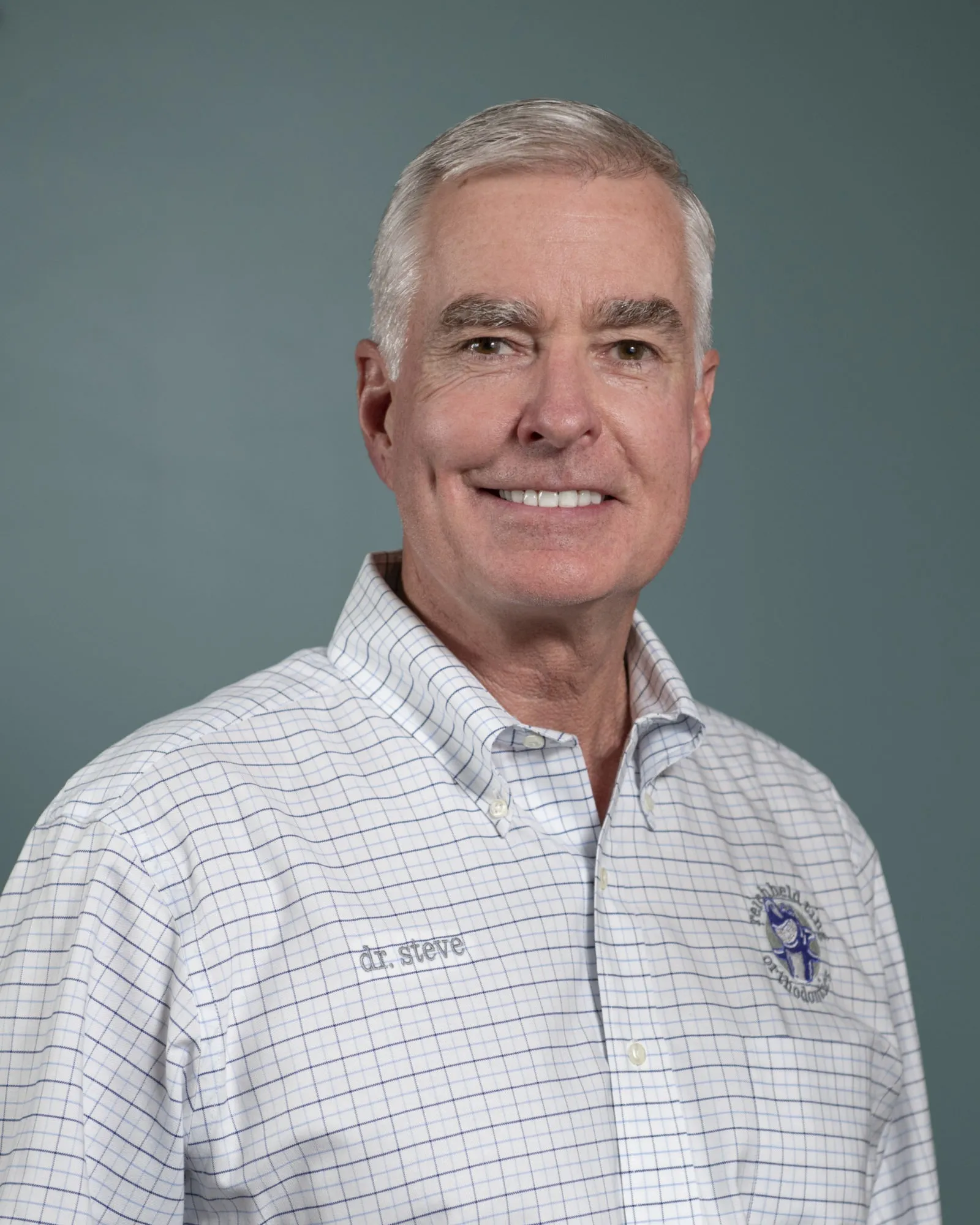 Older man with gray hair smiling wearing a checkered shirt with an embroidered logo and name.