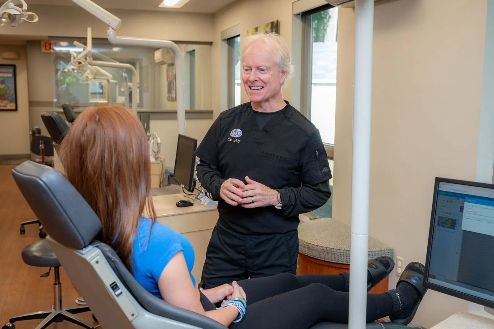 Dentist in black scrubs talking to patient seated in dental chair in bright clinic room.